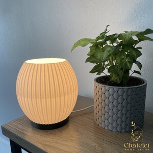 May include: A beige, ribbed, spherical table lamp with a black base sits on a wooden table next to a potted plant. The lamp emits a soft, warm glow. The plant is in a gray, textured pot.