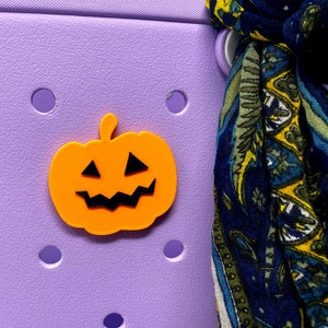 May include: A bright orange pumpkin-shaped charm with a black jack-o'-lantern face. The charm is attached to a purple background.