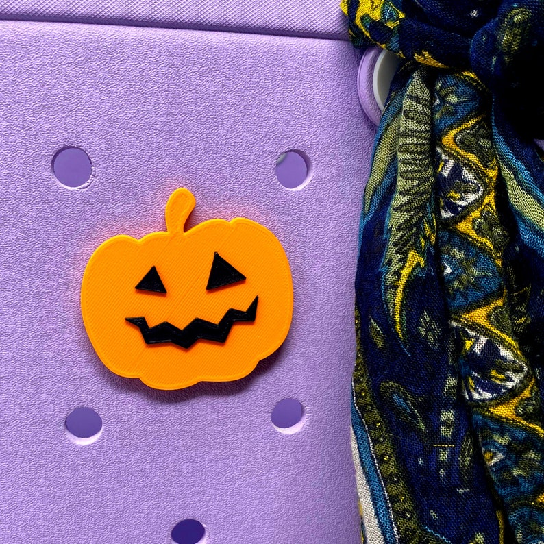 May include: A bright orange pumpkin-shaped charm with a black jack-o'-lantern face. The charm is attached to a purple background.