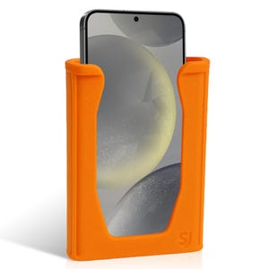 K&ouml;nnte beinhalten: Hellorangefarbener Telefonhalter mit einem Smartphone darin. Der Halter hat ein U-f&ouml;rmiges Design mit abgerundeten Kanten und einer strukturierten Oberfl&auml;che. Der Bildschirm des Telefons zeigt ein unscharfes Bild. Die Initialen "SJ" sind unten rechts sichtbar.