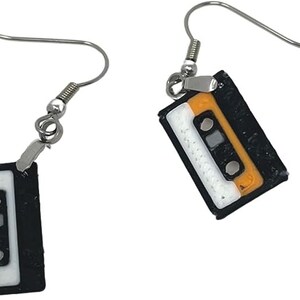 Puede incluir: Un par de pendientes de plata con dijes de cinta de casete negra y naranja. Los dijes tienen una etiqueta blanca con una l&iacute;nea negra en el medio.