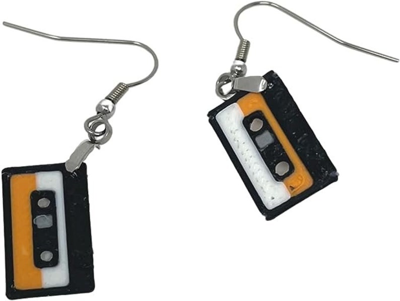 Puede incluir: Un par de pendientes de plata con dijes de cinta de casete negra y naranja. Los dijes tienen una etiqueta blanca con una l&iacute;nea negra en el medio.