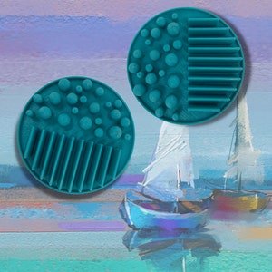 May include: Two teal blue circular scrubbers with raised bumps and ridges. One scrubber has a pattern of raised bumps and the other has a pattern of vertical ridges.