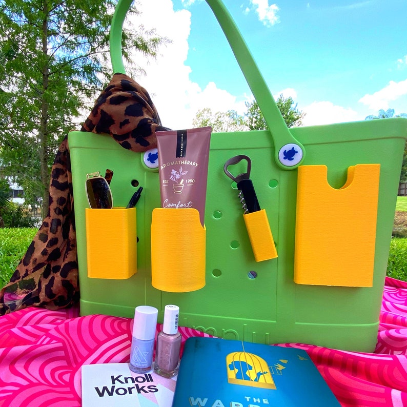 May include: A green and yellow tote bag with various pockets and accessories. The bag has a  "KNOLL WORKS" label and a book with the text "WAR THE" on the cover. The bag is filled with items such as a bottle of lotion, a bottle of nail polish, a bottle opener, and sunglasses.