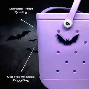 May include: A purple Bogg Bag with a black bat charm and a white clip with the letter "B" on it. The bag has a textured surface with small holes. The text "Durable - High Quality" is visible in the top left corner of the image. The text "Clip Fits All-Sizes Bogg Bag" is visible in the bottom left corner of the image.