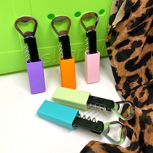 May include: Four colourful bottle openers with a black corkscrew and bottle opener. The bottle openers are arranged in a row on a green surface.