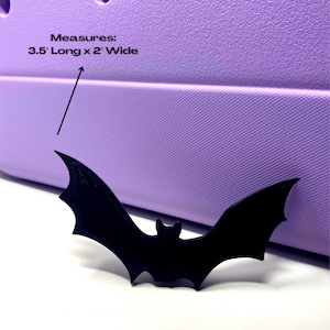 May include: A black bat cutout, 3.5 inches long and 2 inches wide, is placed on a purple surface. The text "Measures: 3.5" Long x 2" Wide" is visible above the bat.