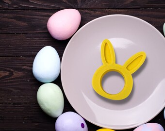 Puede incluir: Un plato blanco con un anillo de servilleta en forma de conejo amarillo en la parte superior. El plato está rodeado de huevos de Pascua de colores pastel.