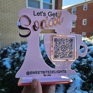 May include: A pink acrylic sign in the shape of a mixer with the words "Let's Get Social" in gold lettering. It includes a QR code and the text "@SWEETBITEDELIGHTS By Lety". The sign is held in front of a snowy background.