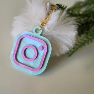 May include: A light blue and pink Instagram logo keychain with a white fluffy pom-pom attached.