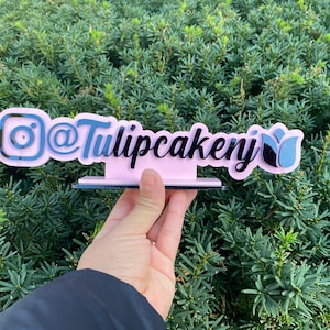 May include: A pink acrylic sign with black lettering that reads "@Tulipcakery" with an Instagram logo and a tulip graphic.