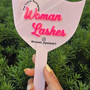 May include: Pink hand mirror with a lip shape. The mirror has the text "Woman Lashes" in pink lettering and the text "Por Laura Rodriguez" in black lettering. The mirror also has the text "Woman_eyelash1" and an Instagram icon.