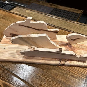 May include: Wooden fish-shaped cutouts arranged on a rectangular wooden board. The fish are various sizes and shapes, with natural wood grain patterns. The board is made of light-colored wood, and the fish are a lighter shade.