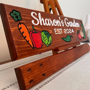 May include: A wooden sign with the text "Sharon's Garden EST 2024" painted in white. The sign is decorated with painted vegetables, including a carrot, bell pepper, cabbage, and a chilli pepper.