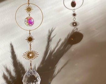 May include: A celestial-themed suncatcher with a gold chain, featuring a faceted crystal ball, iridescent crystal beads, and starburst charms. The suncatcher hangs in front of a wall, casting shadows.