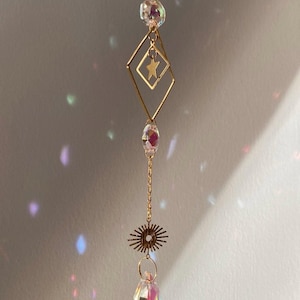 May include: A gold-coloured suncatcher with a chain. It features a diamond-shaped frame with a star, a crystal bead, and a teardrop-shaped prism. The prism reflects a rainbow of colours.