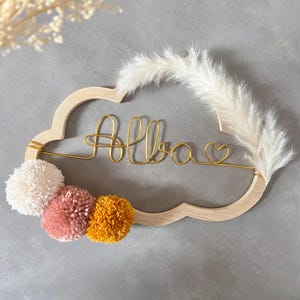 May include: Wooden cloud-shaped wall decoration with the name "Alba" in gold wire. Includes three pom-poms in cream, pink, and yellow, and a fluffy white feather accent. Ideal for a nursery or child's room.