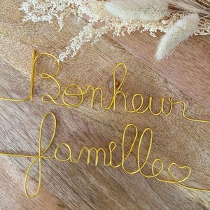 Peut inclure: Décoration murale en fil d'or avec l'inscription "Bonheur famille". Les lettres sont en cursive, avec un cœur à la fin de "famille". Des fleurs séchées ornent l'arrière-plan.