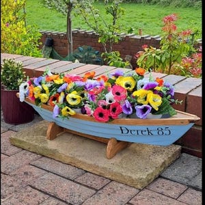 May include: A light blue wooden boat planter filled with colourful flowers, including red, yellow, purple, and white pansies. The boat is supported by wooden legs and sits on a stone base. The text "Derek 85" is painted on the side.