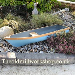 May include: A light blue wooden boat with a grey interior and a wooden seat. The boat has a light brown trim. The boat is displayed on a bed of pebbles and rocks. The website address is visible.