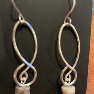 Puede incluir: Un par de pendientes colgantes plateados. Cada pendiente presenta una forma ovalada alargada con un diseño en bucle y una pequeña pila de cuentas de color claro en la parte inferior. Los pendientes se muestran sobre un fondo oscuro.