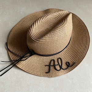 Può includere: Un cappello di paglia marrone con un nastro nero e la parola "Ale" ricamata sul davanti.