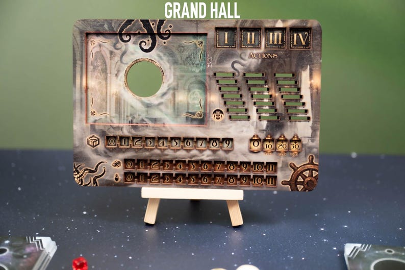 May include: A decorative game board with the text "GRAND HALL". The board features a circular cutout, action trackers, and number indicators. It is supported by a small wooden easel. The color scheme includes shades of gray, brown, and gold.