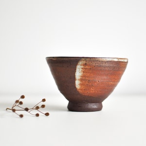May include: A handcrafted ceramic bowl with a footed base. The bowl has a colour gradient, shifting from dark brown to a warm orange-brown hue. A white accent stripe provides contrast. Suitable for serving food, the bowl is approximately 15 cm in diameter.