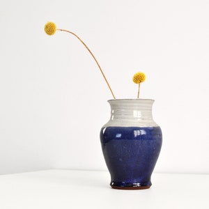 May include: A handcrafted ceramic vase with a two-tone design. The upper section is white, leading to a deep blue base. Two yellow, spherical flowers are placed inside the vase. The vase is approximately 20 cm tall.