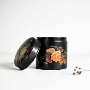 Op de afbeelding: Een zwarte, ronde container met een deksel, versierd met een bloemenontwerp met oranje en gele rozen. Het deksel staat iets open en onthult de binnenkant van de container. Een klein takje gedroogde bloemen ligt in de buurt.