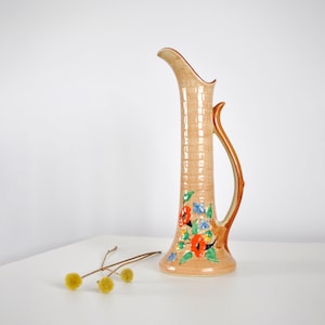May include: A tall, peach-coloured ceramic jug with a woven texture and a curved handle. The jug is decorated with a floral design featuring red, blue, and yellow flowers. A sprig of yellow flowers rests on the white surface.