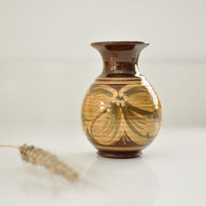 May include: A small, brown ceramic vase with a rounded body and a flared rim. The vase features a floral design in shades of tan and green. A dried plant stem lies in the foreground. The vase is approximately 15 cm tall.
