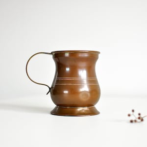 May include: A copper-colored mug with a curved handle. The mug has a wide base and a slightly flared top, with two horizontal lines etched around the middle. The handle is a lighter, brassy color. A small sprig of berries sits to the right.
