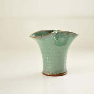 May include: A small, handmade ceramic vase with a flared, scalloped rim. The vase has a light green glaze with darker green and brown accents. The base is a darker brown color. The vase is approximately 6 inches tall.