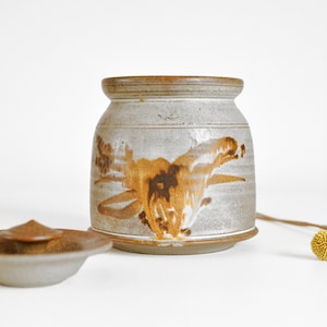 May include: A handmade ceramic jar and lid, in grey and brown tones. The jar is cylindrical with a flared rim and a brown floral design. The lid is a small, shallow dish with a conical knob. The jar and lid are suitable for storage.