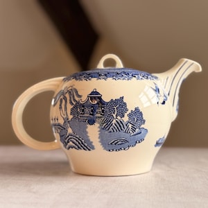 May include: Cream-coloured ceramic teapot with a blue Willow pattern design. The teapot has a curved handle, a spout, and a lid with a small loop. The blue pattern depicts a landscape with trees, buildings, and a bridge.