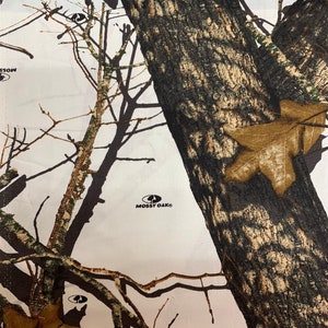 May include: White fabric with a camouflage pattern featuring brown, black, and green branches and leaves. The fabric has the words "Mossy Oak" printed in black.