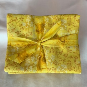 May include: A stack of folded yellow fabric featuring a repeating pattern of small white and dark yellow flowers and thin stems. The fabric is tied together with a matching yellow ribbon that is knotted in the center of the square bundle.