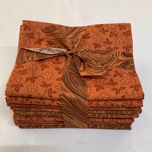 May include: A stack of 10 orange and brown fabric squares with a brown and orange swirl pattern fabric tied around the stack.