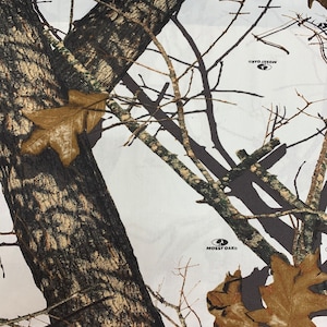 May include: White camouflage fabric with brown and green tree branches and leaves. The fabric has the Mossy Oak logo printed on it.