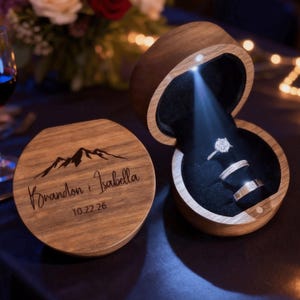 Pode incluir: Uma caixa de anel de madeira com um design de montanha e os nomes "Brandon & Isabella" e a data "10.22.26" gravados na tampa. A caixa está aberta, revelando dois anéis iluminados por uma pequena luz. A caixa está em uma superfície escura.