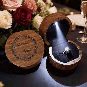 May include: A wooden ring box with a light shining on a diamond ring. The box is open, revealing the ring on a black velvet interior. The lid of the box is engraved with the names "David & Annie" and the date "20.08.26".