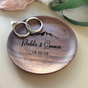 Może przedstawiać: Drewniana podstawka na obrączki z wzorem sylwetki g&oacute;r, spersonalizowana z imionami "Pablo & Emma" i datą "19-10-19". Na podstawce leżą dwa złote obrączki.