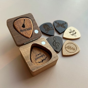 May include: A wooden box with a hinged lid, containing a guitar pick with the text "Edward 19.06.73" inside. The box is open and next to it are five guitar picks with various designs, including a skull, a treble clef, a guitar, a butterfly, and a letter "P".