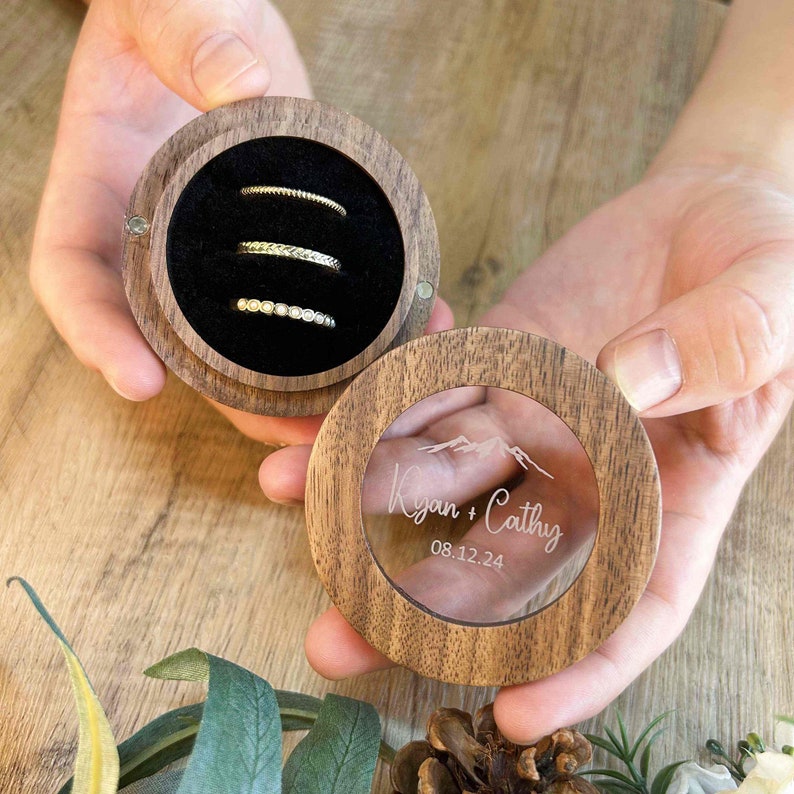 K&ouml;nnte beinhalten: Eine Holzkiste f&uuml;r Ringe mit einem durchsichtigen Deckel. Der Deckel ist mit einem eingravierten Motiv von Bergen und dem Text "Ryan + Cathy 08.12.24" verziert. Die Kiste ist ge&ouml;ffnet und zeigt drei Ringe im Inneren.