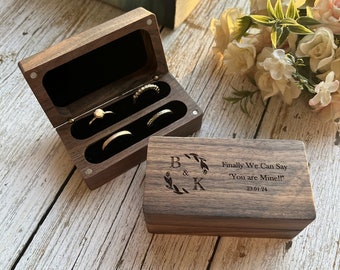 May include: A wooden ring box with a hinged lid. The box has two compartments for rings. The lid is engraved with the letters "B & K" and the text "Finally We Can Say 'You are Mine!!' 23.01.24".