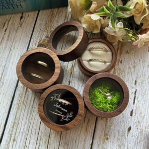 K&ouml;nnte beinhalten: Vier h&ouml;lzerne Ringboxen mit durchsichtigen Deckeln. Zwei Boxen enthalten Ringe, eine Box enth&auml;lt eine Perle und Moos, und eine Box hat eine gravierte Inschrift mit dem Text "A&P Johnson 23.10.23".