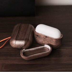 May include: Three wooden cases for wireless earbuds. The cases are made of dark brown walnut wood and have a smooth, polished finish. The cases are open and show the interior of the cases, which are lined with a soft, white material. The cases are arranged in a row on a wooden surface.