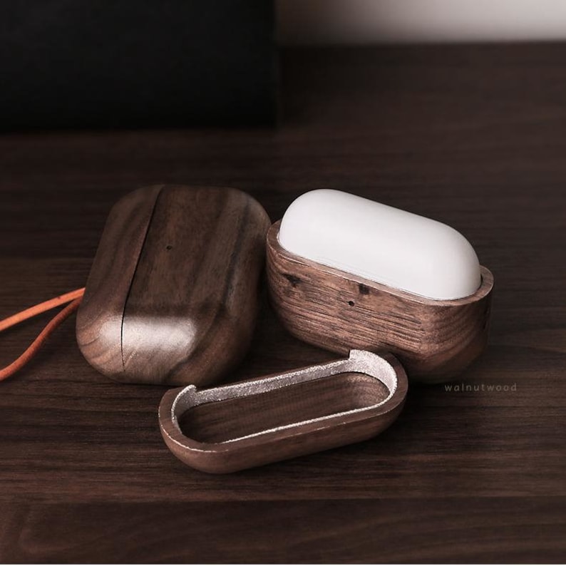 May include: Three wooden cases for wireless earbuds. The cases are made of dark brown walnut wood and have a smooth, polished finish. The cases are open and show the interior of the cases, which are lined with a soft, white material. The cases are arranged in a row on a wooden surface.