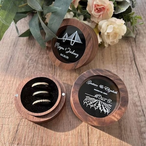 K&ouml;nnte beinhalten: Zwei Holzkisten f&uuml;r Ringe mit schwarzen Glaseins&auml;tzen. Die obere Box hat ein Br&uuml;ckenmotiv und den Text "Megan & Zachary 04.26.25". Die untere Box hat ein Weinbergsmotiv und den Text "Gustavo & Elizabeth MARCH 20, 2023".